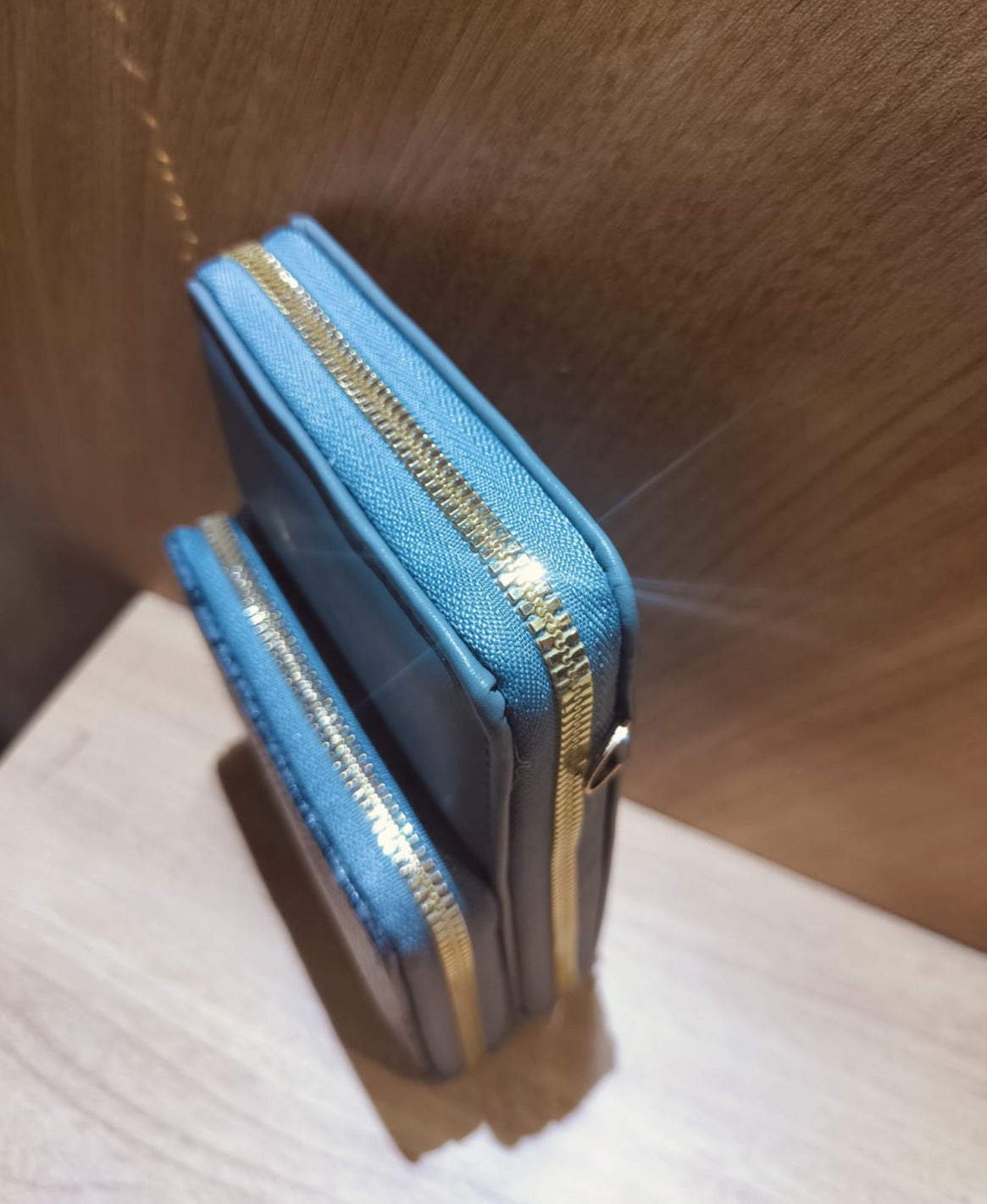 Blue mobile purse sling with gold zipper detail standing against wooden background