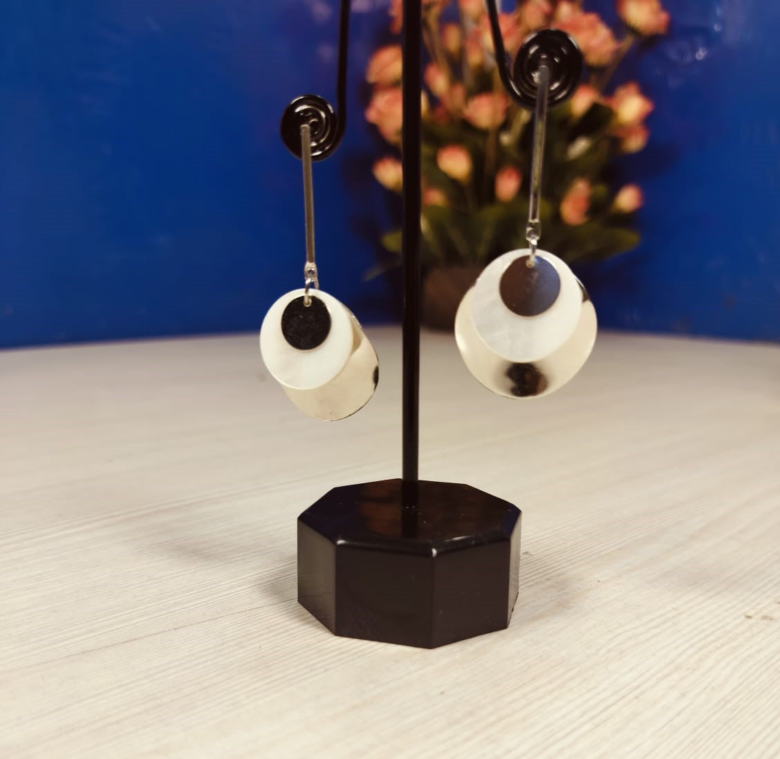 Wincham Fashion House silver and gold seashell earrings displayed on black stand with blurred floral background