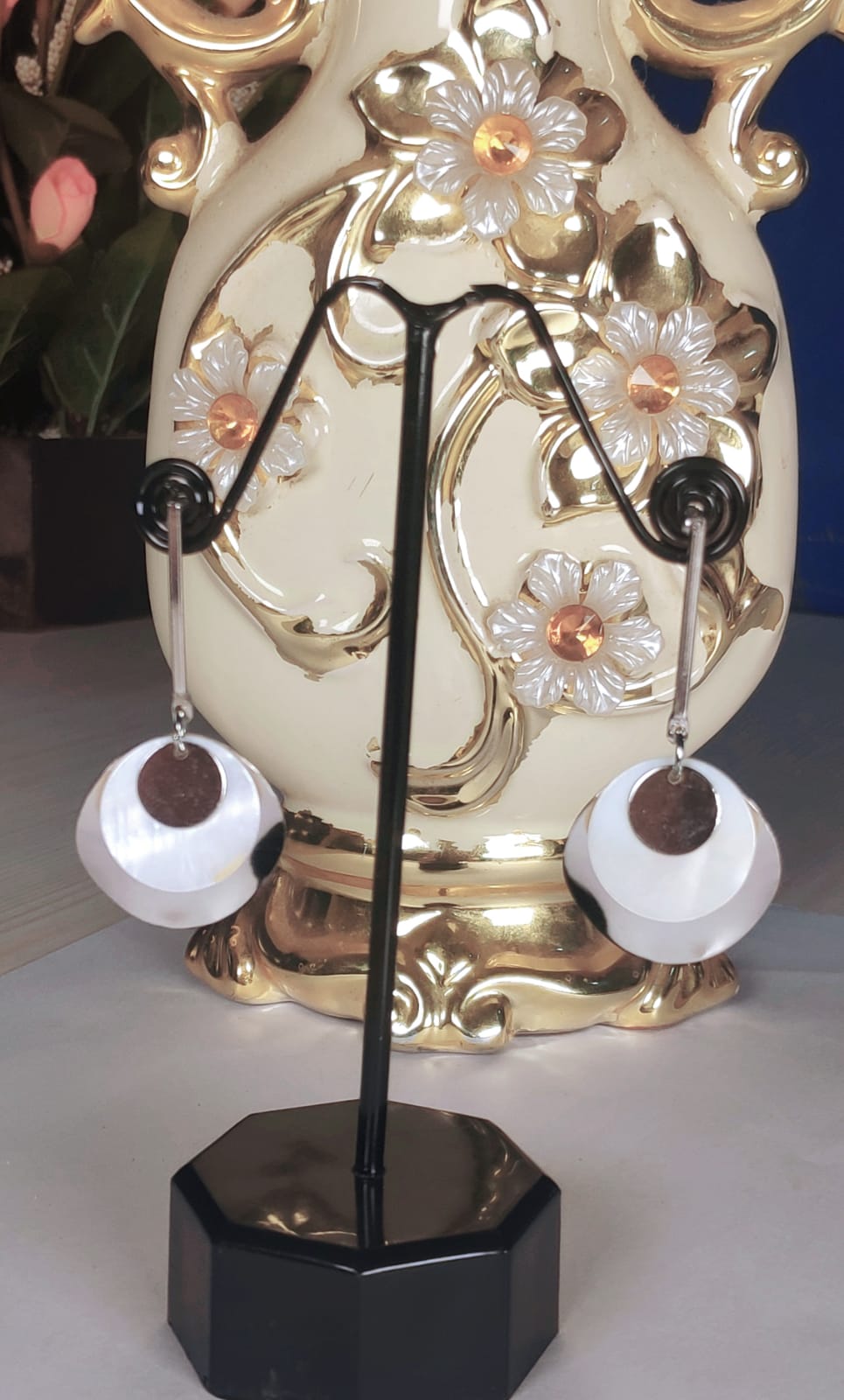 Silver and gold seashell earrings displayed on black stand with decorative gold floral vase background