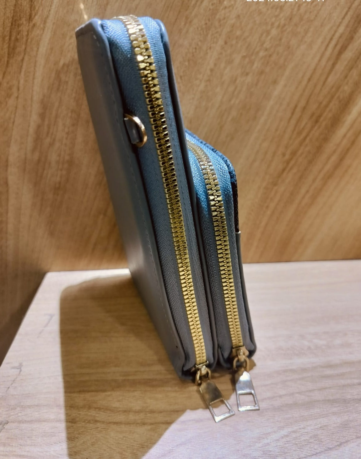 Gray mobile purse sling bag with double gold zippers standing on wooden surface